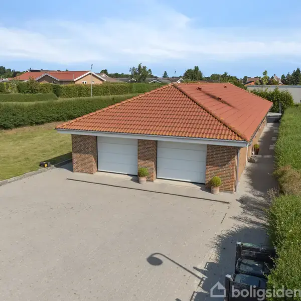 Garagebygning med rødt tegltag og to hvide porte. Den står på en stor asfalteret plads, omgivet af grønne hække og græsarealer i et boligområde.