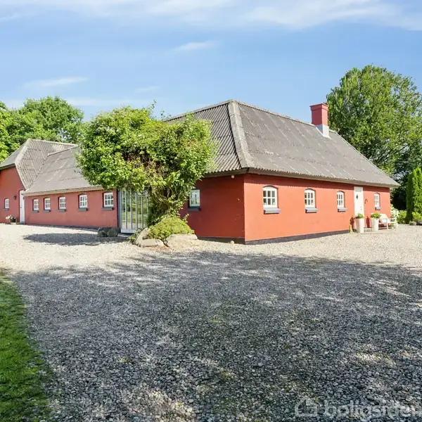 Rødt landhus med hvidrammede vinduer står stille på en grusvej, omgivet af grønne træer og buske under en blå himmel.