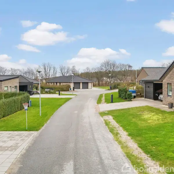 Flere parcelhuse i moderne stil står langs en stille vej. Området er velholdt med grønne plæner og flisebelagte fortove. Skyfri himmel og baggrund af træer skaber en fredelig atmosfære.