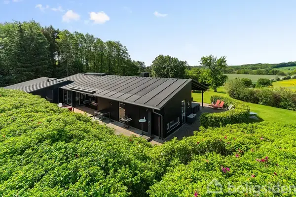Et sortmalet hus med fladt tag er omgivet af frodig hæk og have. Det sidder fredeligt i landskab med skove og marker, der strækker sig i baggrunden under den blå himmel.