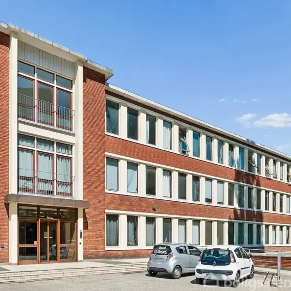 En tre-etagers bygning af rød mursten har adskillige store vinduer. Nogle biler er parkeret foran bygningen. Omkring er der et åbent område med blå himmel og et træ ved siden.