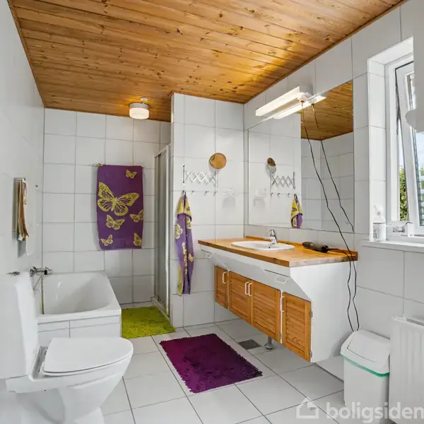 Et badeværelse med hvidt flisegulv og træloft. Et badekar og toilet er til venstre. En vask med spejl er til højre, flankeret af dekorative håndklæder. En vindue giver naturligt lys.