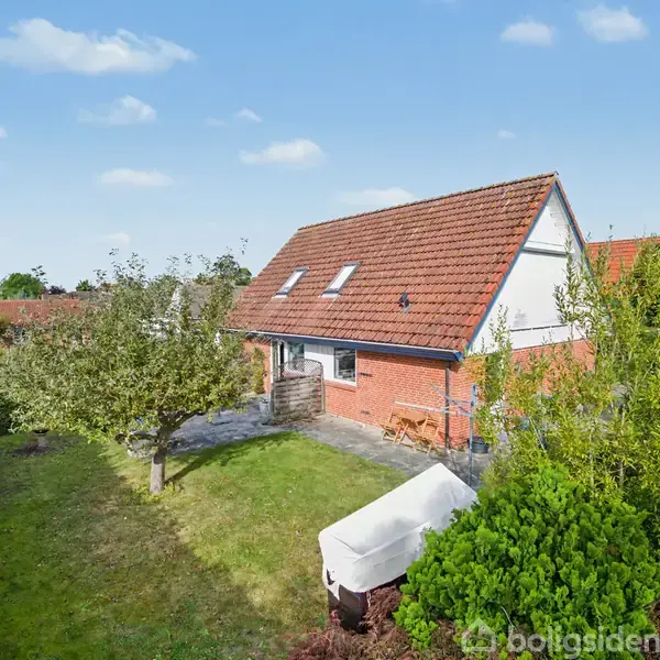 Rødt murstenshus med rødt tegltag, omgivet af grøn have med træer og buske. En terrasse er foran huset, og himlen er blå med få skyer.