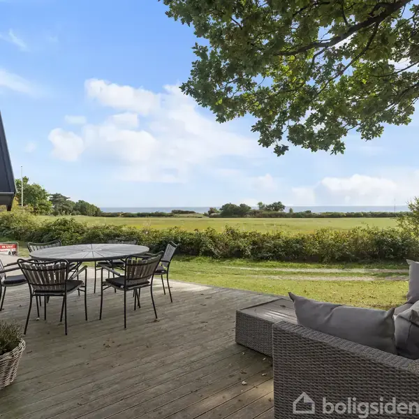 En udendørs terrasse med en rund spisebord og stole står på en trædæk, omgivet af grønne buske og udsigt til marker under en skyfri blå himmel.