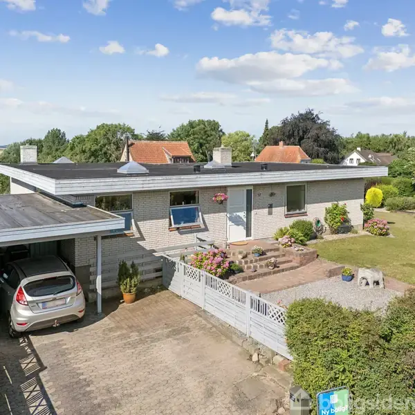 Et enplanshus med fladt tag står i en velplejet have. En bil er parkeret i en carport på venstre side. Blomsterkasser pynter ved huset. Gård belagt med fliser.