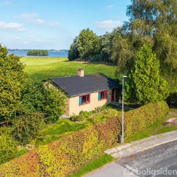 Et hus omgivet af frodig vegetation og en vej ses i forgrunden. Huset har et mørkt tag og skorsten, beliggende i et landligt område med udsigt til en sø i baggrunden.