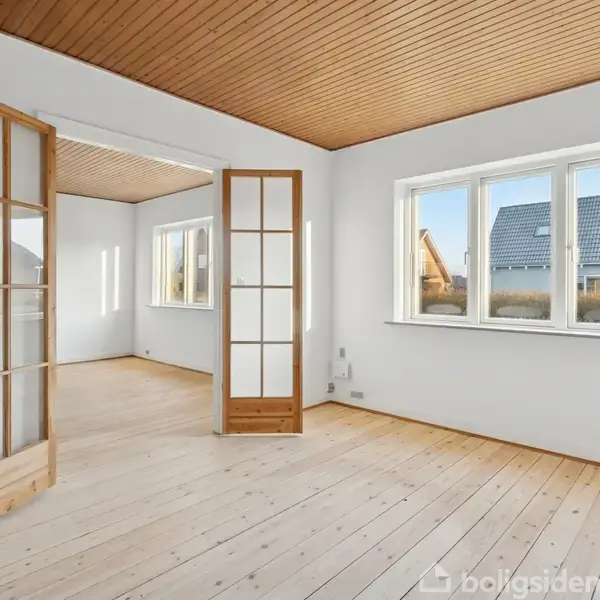 Et tomt rum med trægulv og træloft har en glasdør stående åben, der fører til et tilstødende rum. Store vinduer fylder rummet med naturligt lys.