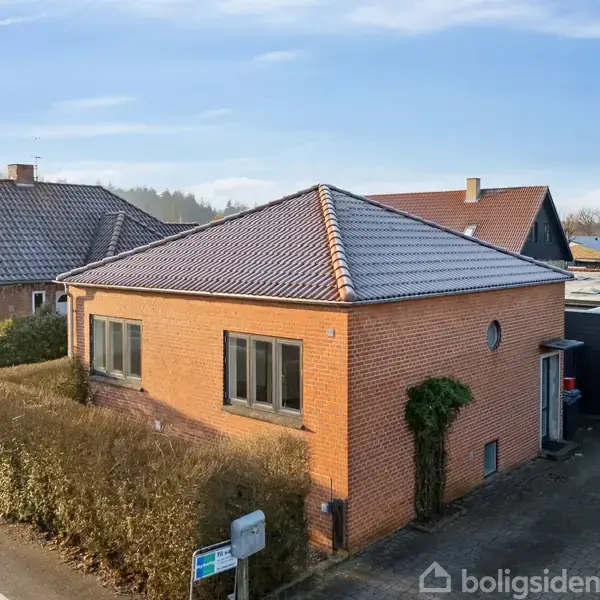 Rødt murstenshus med mørkt tegltag står ved en vej med en lav hæk omkring. I baggrunden ses en anden bygning og nogle træer under en blå himmel.