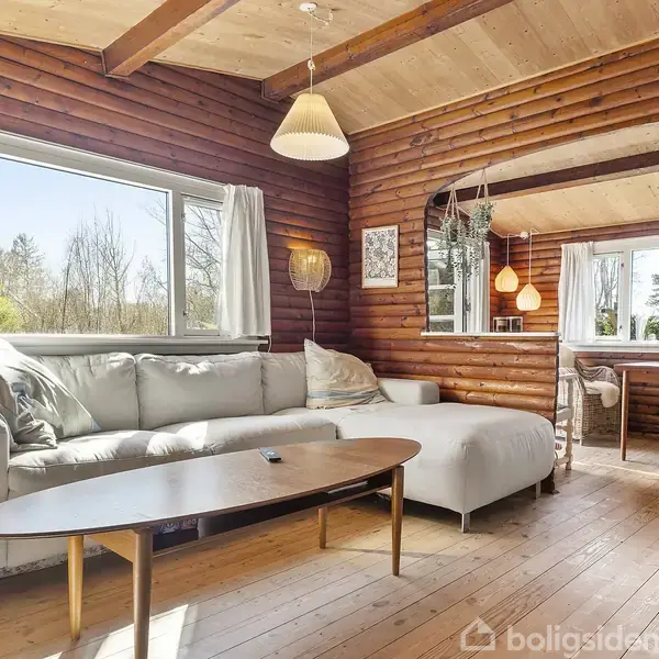 En lang, hvid sofa står i en stue med trævægge og trægulve. Sofaen er placeret foran et stort vindue med udsigt til træer. En lys træbord er foran sofaen.