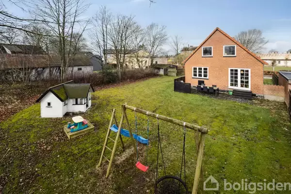 Et rødt murstenshus med hvide vinduer står i en have med gyngestativ, legehus og bord. Træer omkranser græsplænen.