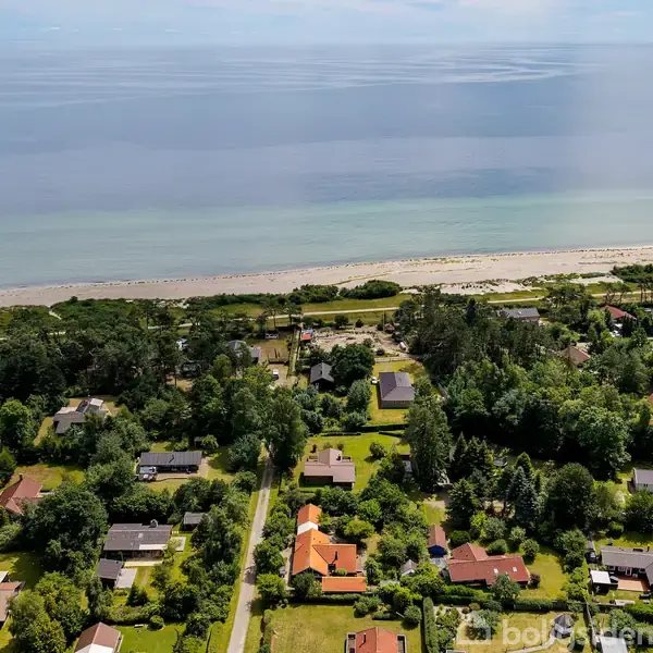 Kystnært boligområde med huse spredt blandt træer og haver. En vej løber gennem byen, og i baggrunden ses en strandlinje og havet.