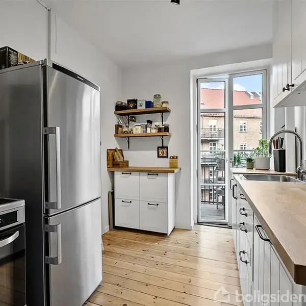 Køleskab står ved siden af en komfur i et køkken med trægulv; køkkenbordet har en skål med citrusfrugter. En dør fører ud til en balkon med udsigt til bygninger.