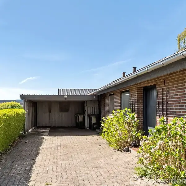 En murstensbygning med hække på den ene side, en carport til venstre og et flisebelagt område i forgrunden. Sollys oplyser scenen, hvilket skaber et lyst miljø.