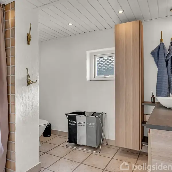 Et rummeligt badeværelse med bruser til venstre, en vask med spejl til højre og en vasketøjskurv i midten. Væggene er lyse, og der er et vindue over kurven.