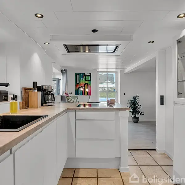 Et moderne køkken med hvide skabe og sort vask, omgivet af lyse fliser. Et stort vindue lader dagslys ind. Der står en plante og kunstværk i baggrunden.