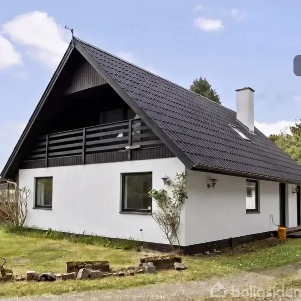 Et hvidt, trekantet hus står stille i en landlig have. Taget er sort, og der er en altan. Buske omgiver huset. Tekst "Visualisering" vises øverst til højre.