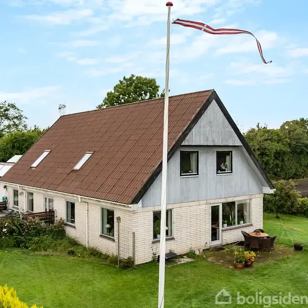 Et to-etagers hus med rød-brunt tag står omgivet af grønne buske og træer. En flagstang med flagrende vimpel er placeret foran huset på en velholdt græsplæne.