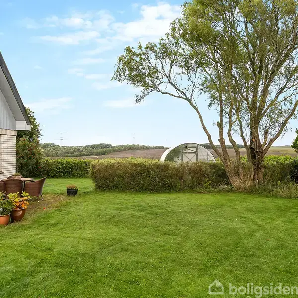 En velplejet græsplæne strækker sig foran et hus med en stor vinduesfacade. Et træ og buske danner en naturlig ramme, mens landskabet breder sig mod horisonten.