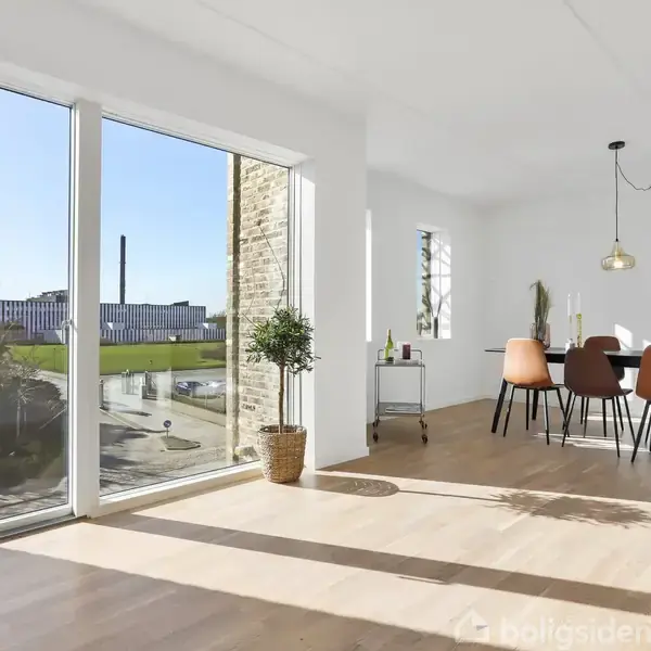 Stue med stort vindue, der giver udsigt til en gårdhave og grønne områder. Spisebord med farverige stole står under en hængelampe. Gennemsigtig flaske står på en vogn.