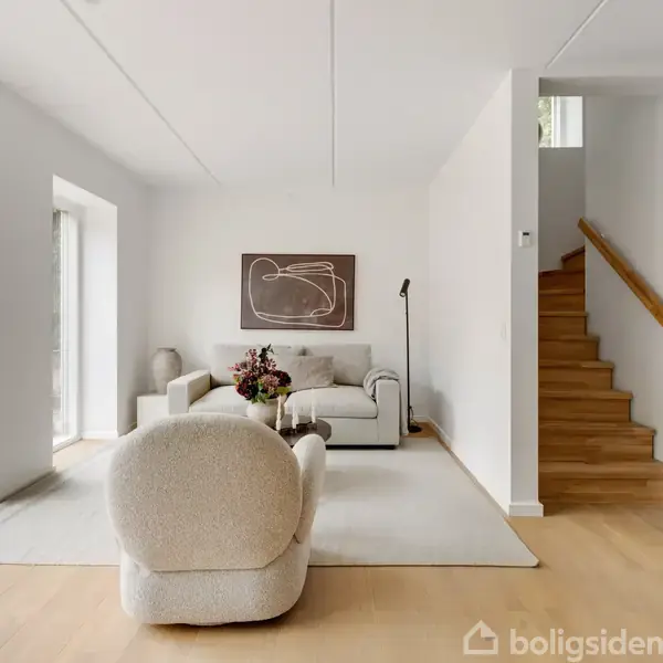 En lys stue med en beige lænestol og sofa på et tæppe. En trappe leder opad til højre. Der hænger abstrakt kunst på væggen, med naturligt lys fra et vindue til venstre.