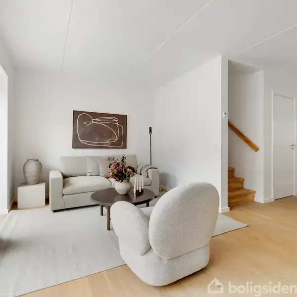 En lys stue med moderne møbler: en sofa og lænestol omkring et sofabord. Væggene er hvide, og der hænger kunst på væggen. Trappe til højre fører til næste etage.