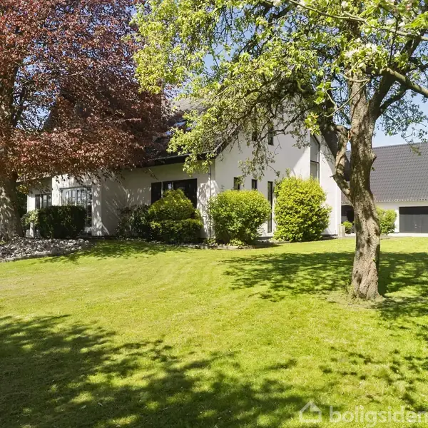 Et hvidt hus står stille omgivet af en velplejet grøn græsplæne og store træer med farverige blade. Solen skinner, hvilket skaber skygger på jorden.