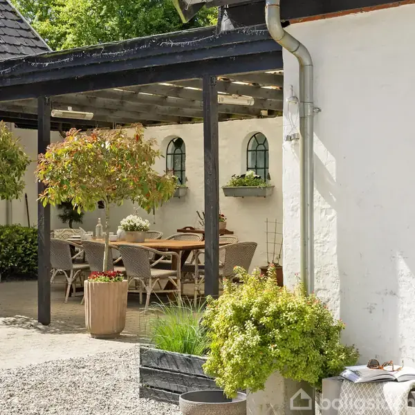 Terrasse med træmøbler under en pergola, omkranset af planter i krukker. En hvid bygning med små vinduer udgør baggrunden, mens grusbelagt jord dækker området.
