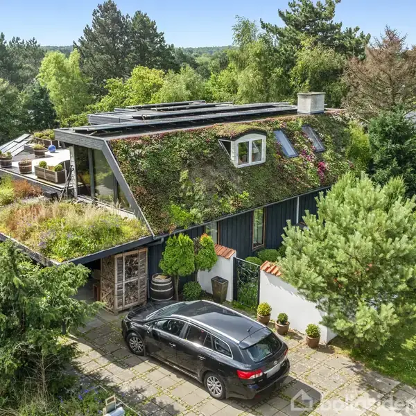 En bil står parkeret foran et moderne hus med et grønt tag og solpaneler. Omkring huset er der frodig vegetation og træer, hvilket skaber en naturrig atmosfære.