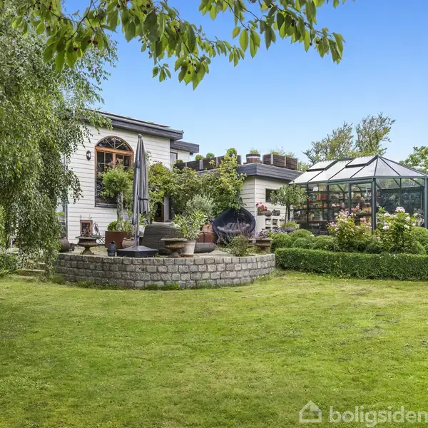Et hus med terrasse står blandt grøn vegetation. Ved siden af er et drivhus omgivet af blomster. Græsplæne og buske udgør haven, mens himlen er klar og blå.