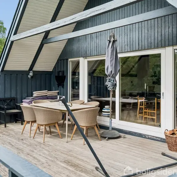 Terrasse med træmøbler og parasol står foran en moderne, mørkeblå træhusfacade med store vinduer. Der er en kurv med planter til højre og grønne træer omkranser området.