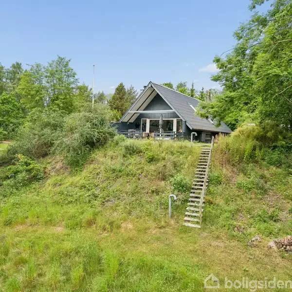 Et sommerhus står på en bakke omringet af frodig skov. En trappe fører op til terrassen foran huset, som har et mørkt tag og store vinduer.
