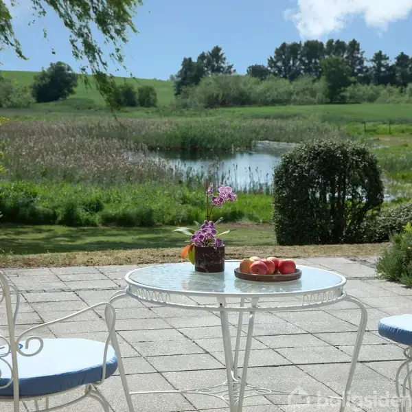 Et rundt bord med blomster og frugt står på en terrasse med udsigt til en sø og grønne marker i baggrunden. To stole med blå hynder står ved bordet.