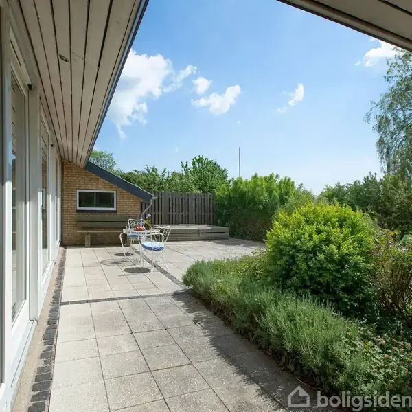 En terrasse med fliser og et ryddeligt haveområde, komplet med frodig vegetation. I baggrunden står et bord med stole. Solrig himmel og et træ er synligt i horisonten.