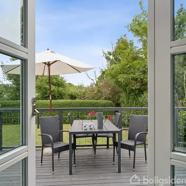 En åbent terrassedør viser et udendørssæt med bord og fire stole på en træterrasse. En parasol står i haven, omgivet af grønne træer og buske.