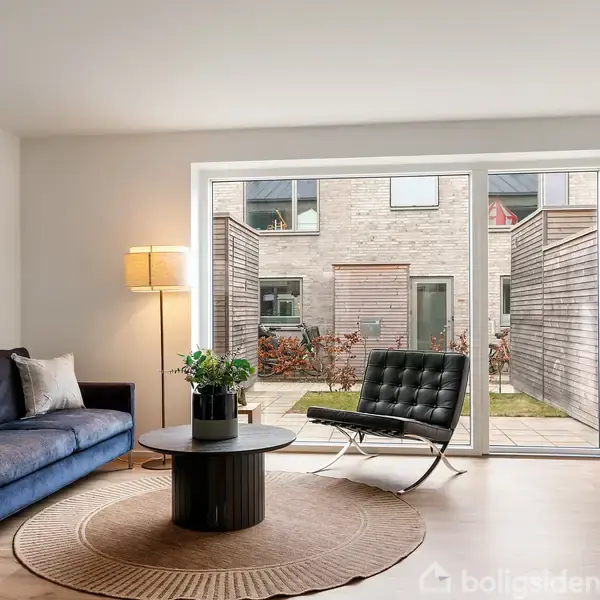 En stue med en blå sofa og sort lænestol står ved et rundt sofabord. Store vinduer viser en gårdhave med moderne byhuse. En lampe står tændt ved sofaen.