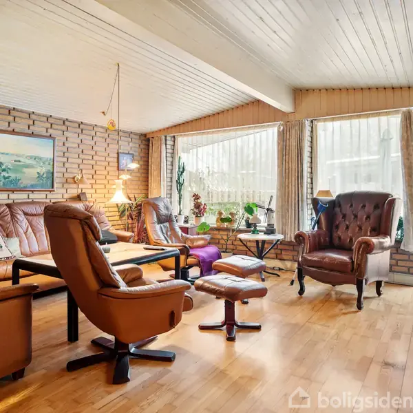 En stue med læderstole står omkring et lavt sofabord. Rummet har træloft, murstensvægge, og store vinduer, der giver udsigt til en snedækket have. En gul stol udenfor.