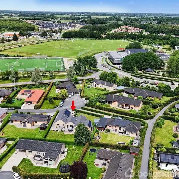 En bolig i et villakvarter er markeret med en rød pil. Omkringliggende huse har haver. I baggrunden ses en stor grøn sportsplads og flere bygninger ved siden af.