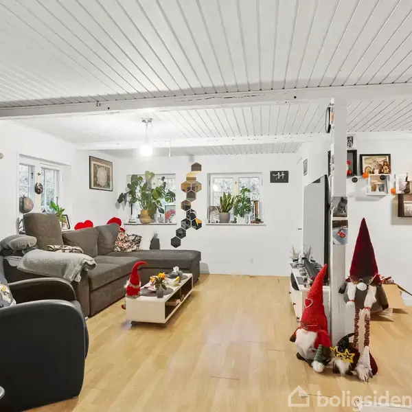 Stue med grå sofa, pyntegenstande og planter i vindueskarmen. På væggen hænger en dartskive. Gulvet er lyst trægulv, og der er julekalkunst. Billedrammer og dekorationer ses også.