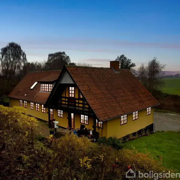 Et gult bindingsværkshus med rødt tegltag er oplyst indefra og står i en landlig omgivelser med marker og træer i baggrunden, under en klar aftenhimmel.