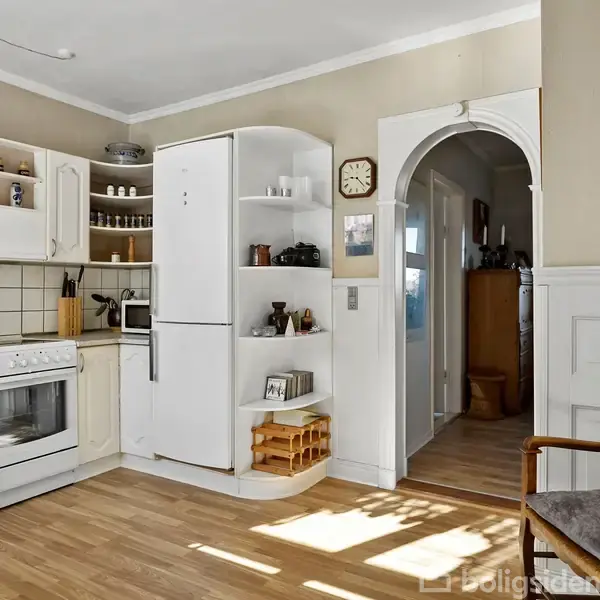 Et køkken med hvide skabe og en ovn ved siden af et stort køleskab. En åbning fører til en gang. Der er trægulv og en stol ved siden af. En vægur hænger på væggen.