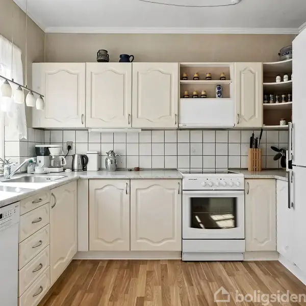 Køkken med lyse skabe og hvidevarer, herunder opvaskemaskine, komfur og køleskab. Skabene indeholder kopper og krukker. Store vinduer fylder rummet med lys. Lyst trægulv og flisebelagt væg.