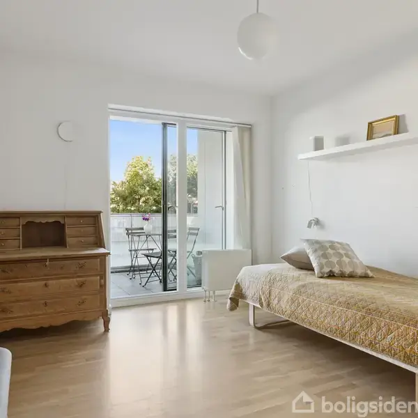 En soveværelse med en enkeltseng dækket af et gyldent tæppe står ved siden af en trækommode. En dør fører ud til en balkon med bord og stole i en lys dagligstue.
