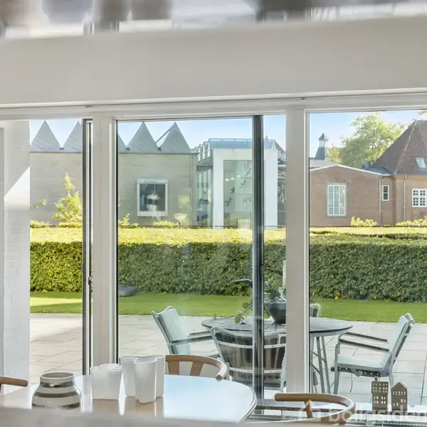 Spisebord med kander og kop foran store glasdøre, der åbner ud til en flisebelagt terrasse. Hækken og det åbne landskab ses i baggrunden, med moderne huse i det fjerne.
