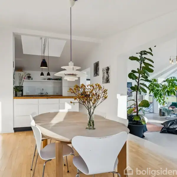 Rundt spisebord står ubevægeligt i en lys stue med trægulv, omgivet af moderne stole, store grønne planter, og overskyet køkken med hvide skabe i baggrunden.