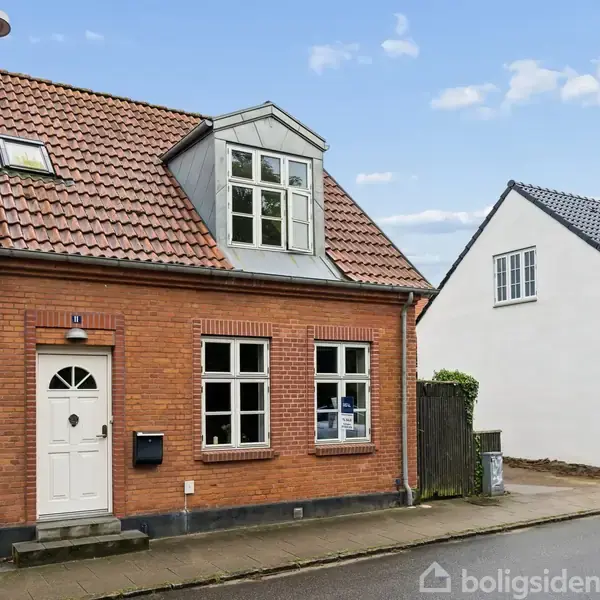 Rødt murstenshus med hvide vinduer og dør står langs en vej. Tagvindue ovenpå og nabohus i hvidt. Skyfri himmel i baggrunden. Skilt ved vindue viser "Bolig til salg".