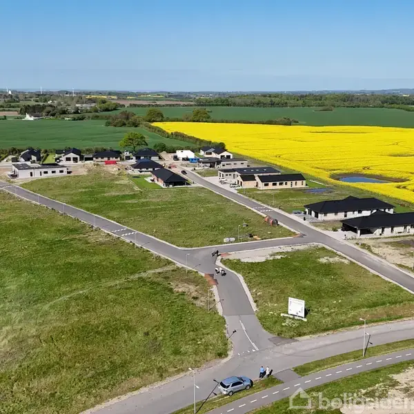 En bebyggelse med nybyggede huse, omgivet af åbne grønne marker og felter af gule blomstrende afgrøder under en klar blå himmel. Veje forbinder strukturerne i et roligt landskab.