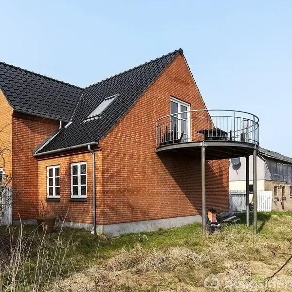 Et rødt murstenshus med sort tegltag har en rund balkon på første sal. Huset står i en åbent område med græs, omgivet af træer og en stor lade i baggrunden.