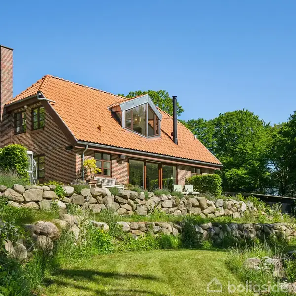 Et rødt murstenshus med rødt tegltag ligger på en bakke omgivet af sten og frodig vegetation. Sollys skinner, og store træer danner baggrund.