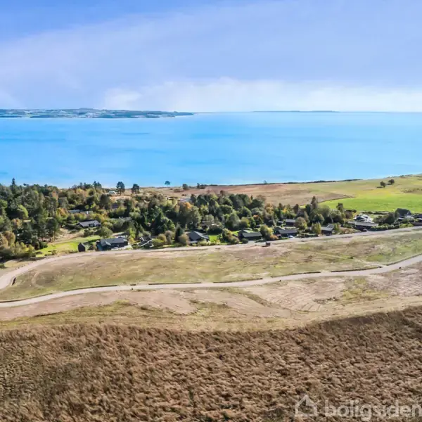 Kystlinje strækker sig langs klart vand; en grusvej løber gennem spredte huse omgivet af marker og skove. Indlands arealer er grønne med rapsmarker under en blå himmel.