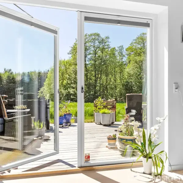 En moderne mørkegrå dør med et diamantformet vindue er omgivet af en pæn gårdhave. Ved siden af står et hvidt bord med planter, dekorativ and og haveudstyr.